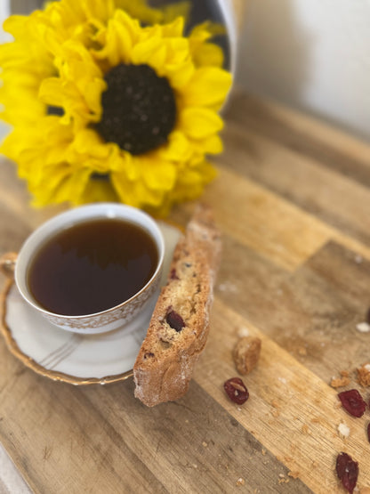Cup of coffee with a biscotti on a wooden surface, accompanied by sunflowers in the background.